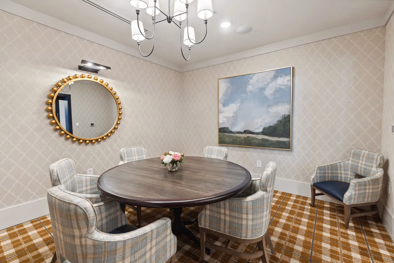 A small dining room with a round wooden table surrounded by six plaid upholstered chairs. A vase with flowers is placed in the center of the table. The room features patterned wallpaper, a large round mirror with a decorative gold frame on one wall, and a landscape painting on another wall. A chandelier with multiple lights hangs from the ceiling, and the floor is covered with a checkered carpet.