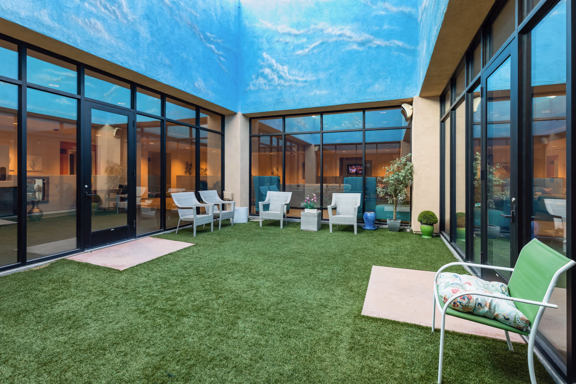 Indoor courtyard area with artificial grass flooring, surrounded by glass walls and doors. Several white chairs and small tables are arranged around the space, along with potted plants. The ceiling is painted with a sky mural featuring clouds.
