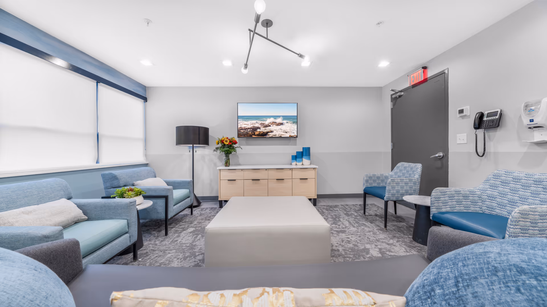 A modern and clean lounge area with multiple blue and gray upholstered chairs and sofas arranged around a central rectangular ottoman. A wooden cabinet with a vase of flowers and decorative items sits against the wall beneath a mounted flat-screen TV displaying an ocean scene. The room has large windows with white blinds, a floor lamp, a gray door with an exit sign above it, and a wall-mounted telephone and hand sanitizer dispenser.