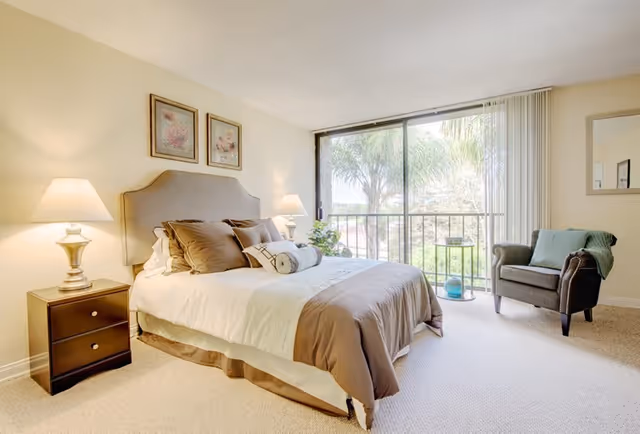 A bright bedroom with a large bed featuring white and brown bedding, two bedside tables with lamps, two framed pictures above the headboard, a gray armchair with a green throw blanket, and a large sliding glass door leading to a balcony with a view of palm trees.