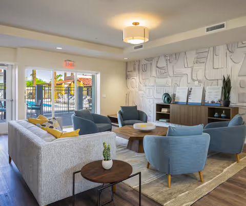 A modern living room in a senior living facility with a light gray sofa adorned with yellow and patterned pillows, four blue armchairs arranged around a wooden coffee table, a small round side table with a small cactus plant, and a wooden cabinet against a textured wall. Large glass doors open to an outdoor patio area with seating and greenery visible outside.