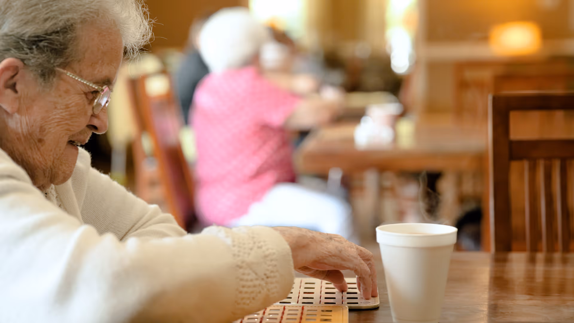 An elderly woman wearing glasses and a white sweater is sitting at a wooden table in a communal dining area, reaching towards a game board. In the background, other elderly individuals are seated at tables, with warm lighting and wooden furniture creating a cozy atmosphere.