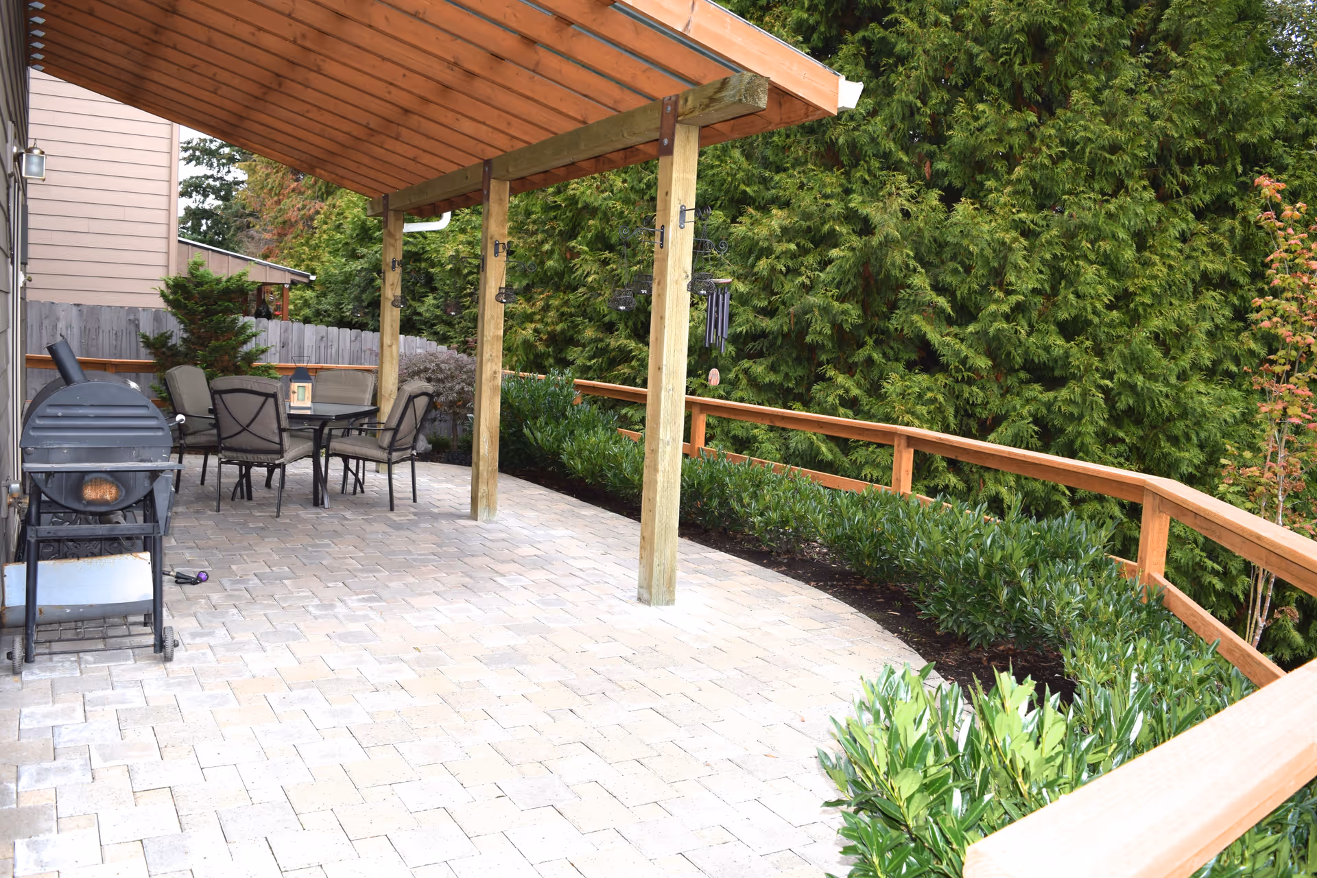 Outdoor patio area with a wooden pergola roof, a black grill, a glass-top table with four cushioned chairs, surrounded by green shrubs and tall trees, with a wooden railing along the edge.