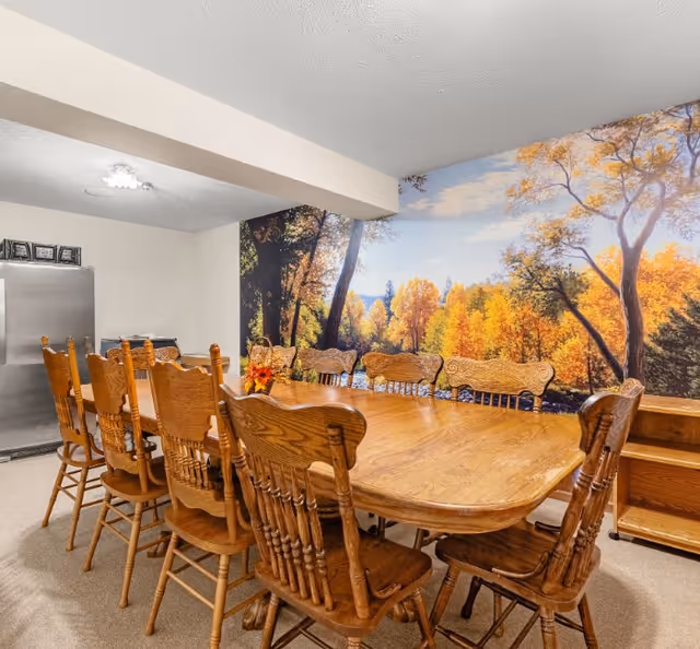 A large wooden dining table surrounded by wooden chairs in a room with a mural of an autumn forest on the wall. The room has beige walls and carpeted floor, with a stainless steel refrigerator in the corner.