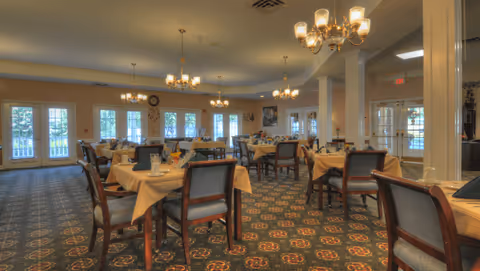 A spacious dining room with multiple tables covered in beige tablecloths, each set with glasses, napkins, and silverware. The room features patterned carpet, several chandeliers hanging from the ceiling, large windows letting in natural light, and a door leading outside.