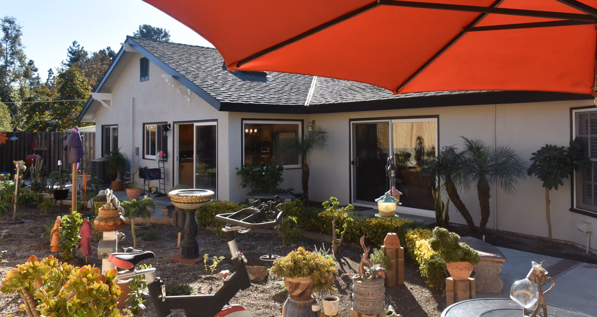 Outdoor garden area with various potted plants, garden decorations, and a large red umbrella providing shade. The background shows a single-story building with multiple windows and sliding glass doors.