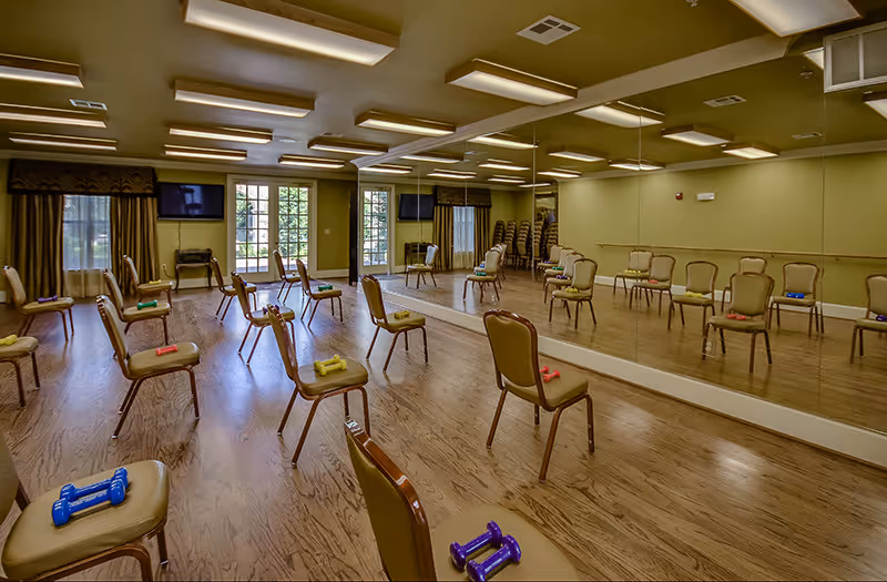 A spacious exercise room with wooden floors and green walls, featuring multiple chairs arranged in rows with colorful dumbbells placed on each chair. The room has large mirrors on one wall, two flat-screen TVs mounted on the opposite wall, and windows with curtains allowing natural light to enter.