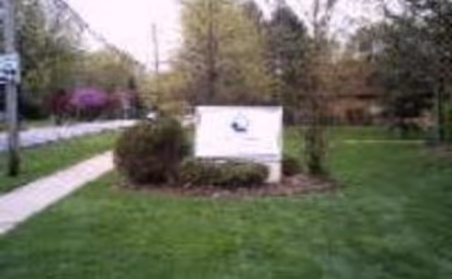 A grassy outdoor area with a sidewalk on the left side and a white sign in the center surrounded by bushes and trees in the background.