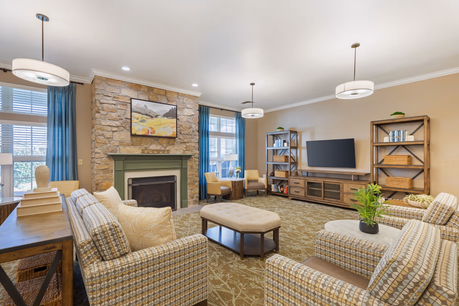 Bright communal living room with a stone fireplace, patterned armchairs, an ottoman, a TV on a wooden console and bookshelves.