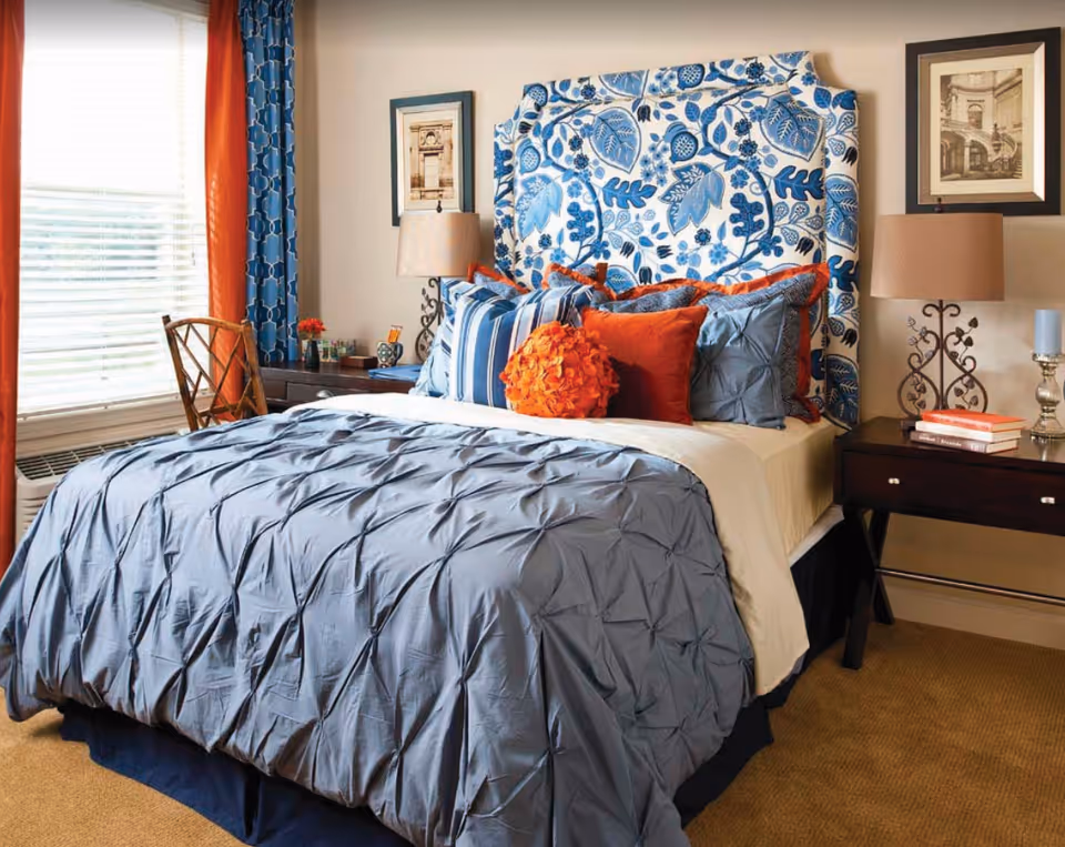 A bedroom with a bed featuring a blue and white floral patterned headboard, blue and orange pillows, and a blue comforter. There are two dark wooden nightstands on either side of the bed, each with a lamp and decorative items. A window with blue and orange curtains is on the left side of the room, and a wooden chair is positioned near the window.