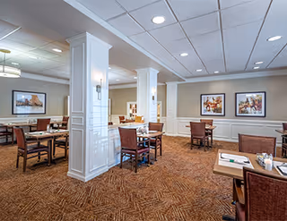 A spacious dining room with multiple wooden tables and chairs arranged neatly. The room features white paneled columns, beige walls with framed artwork, and a patterned carpet. The ceiling has recessed lighting and a grid pattern.