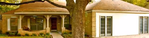 Single-story senior living building with a front porch, large tree, and glass doors.