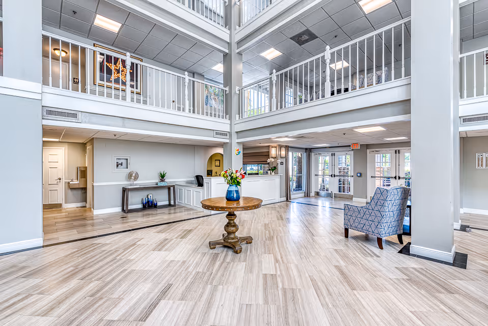 Spacious and bright senior living facility lobby with light wood flooring, a round wooden table with a blue vase of flowers in the center, a blue patterned armchair, white reception desk, and a second-floor balcony with white railings overlooking the area.