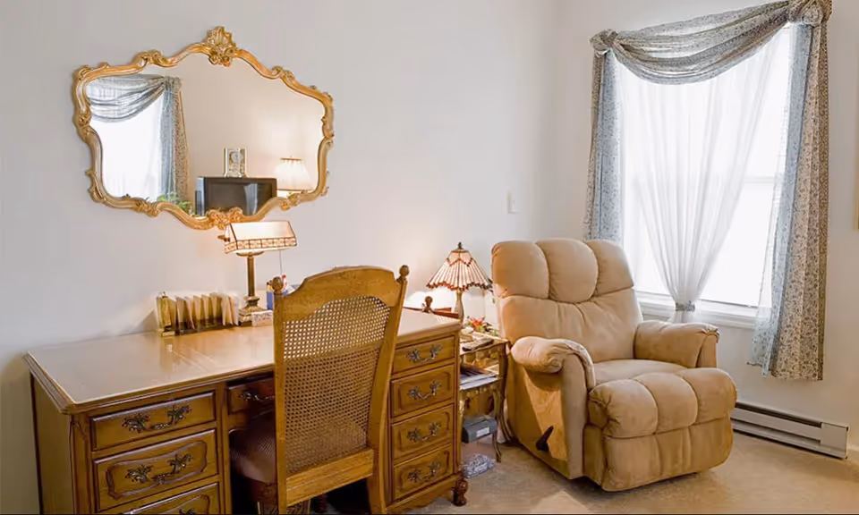 A cozy room featuring a wooden desk with multiple drawers, a matching wooden chair with a woven back, a decorative gold-framed mirror above the desk, a small table lamp on the desk, and a beige recliner chair next to a window with sheer white curtains and floral drapes.
