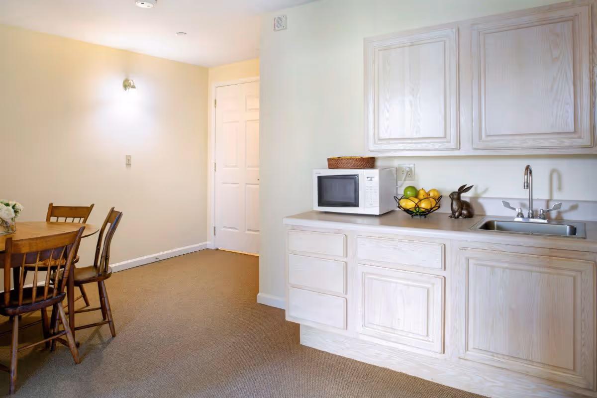 A small kitchen area with light wood cabinets, a white microwave, a fruit basket with lemons and green apples, and a decorative rabbit figurine on the countertop next to a sink. To the left, there is a wooden dining table with four chairs against a beige wall with a wall-mounted light fixture and a closed white door in the background.