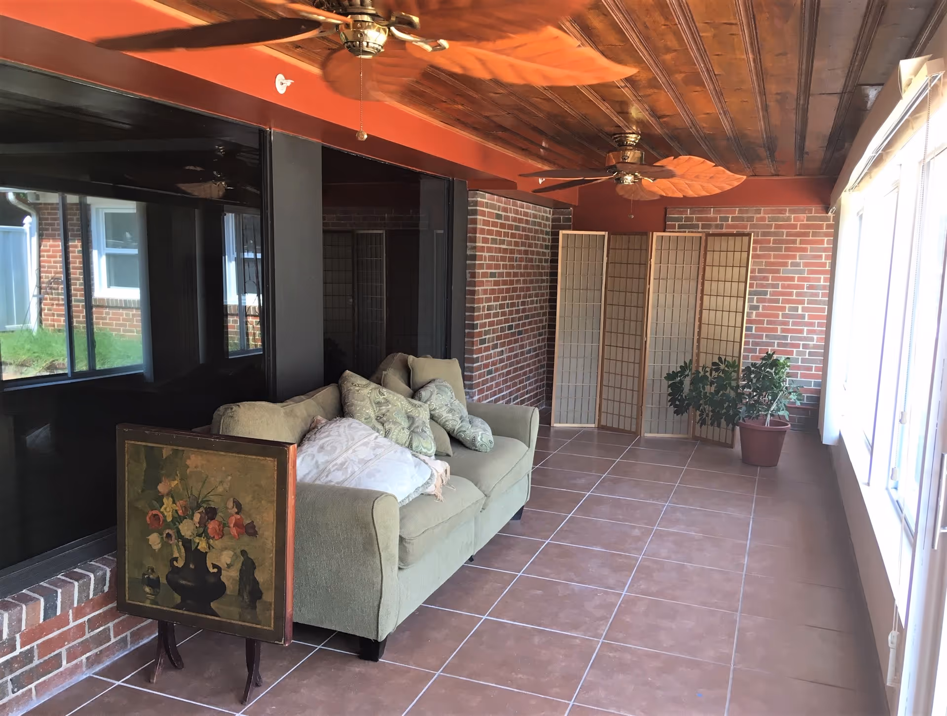 Sunroom-style sitting area with a green sofa and pillows, folding screens, a potted plant, ceiling fans, brick walls and tiled floor beside large windows.