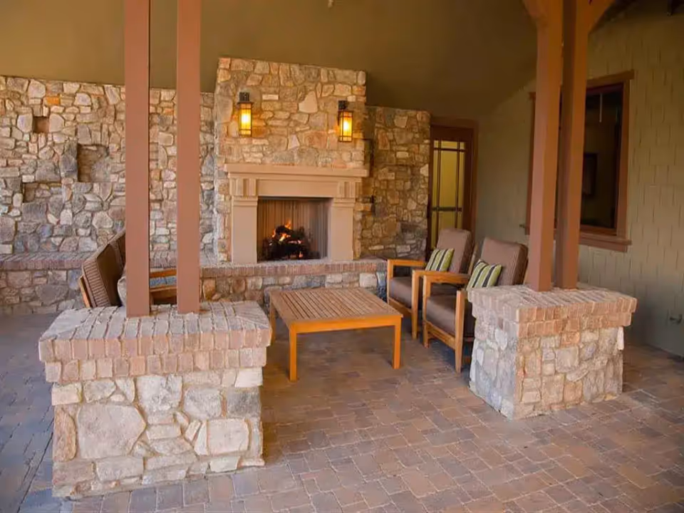 Covered outdoor seating area with stone pillars and walls, featuring a lit fireplace, two cushioned armchairs with striped pillows, and a wooden coffee table on a brick floor.