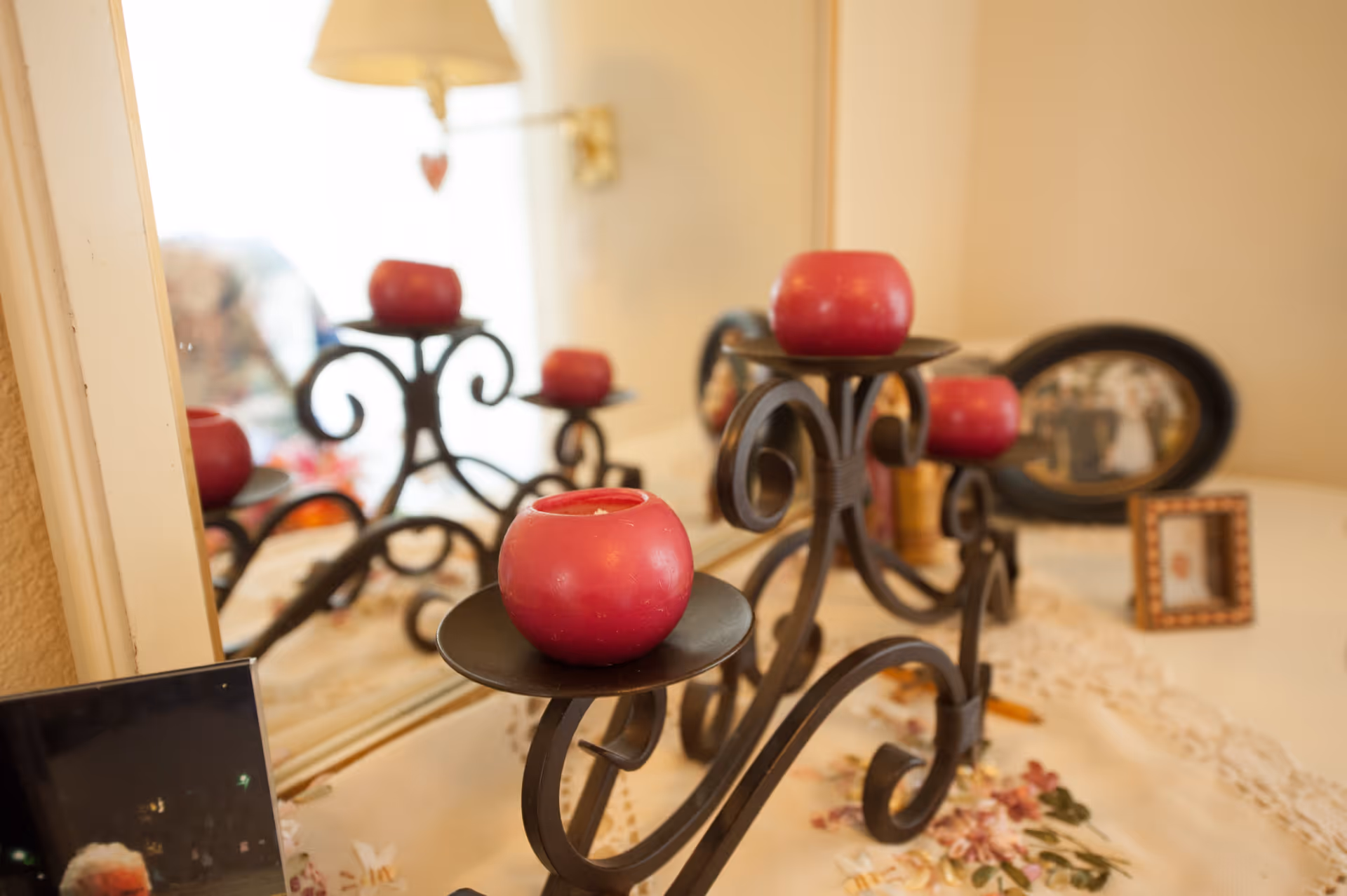 A decorative black metal candle holder with four red candles placed on a white surface with a floral cloth. Behind it is a mirror reflecting the candle holder and candles. There are also framed photos in the background on the surface.