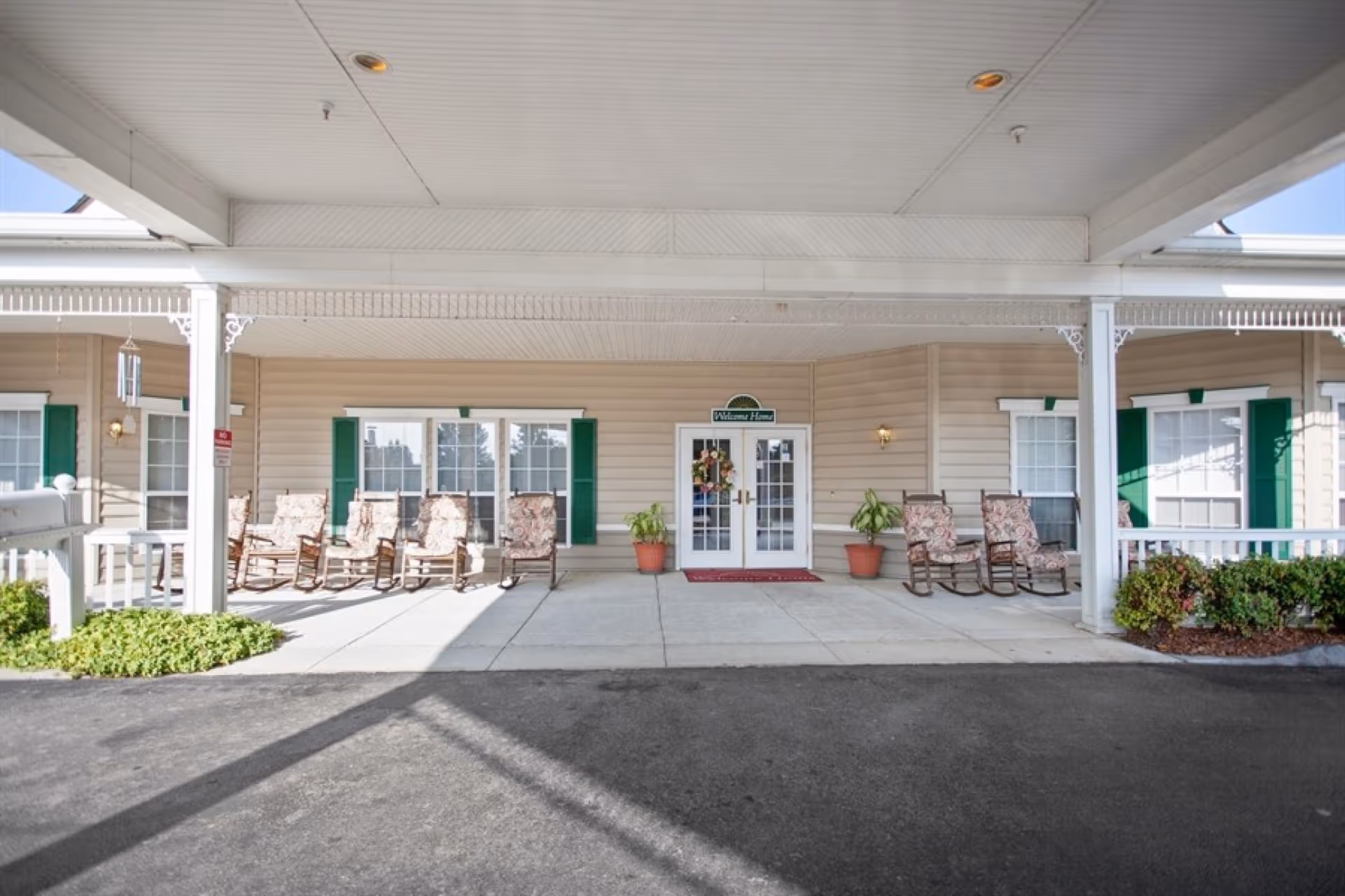 Covered entrance area of a building with a row of cushioned rocking chairs on either side of double glass doors. The doors have a wreath and a sign above them that reads 'Welcome Home'. There are green shutters on the windows and potted plants near the entrance.