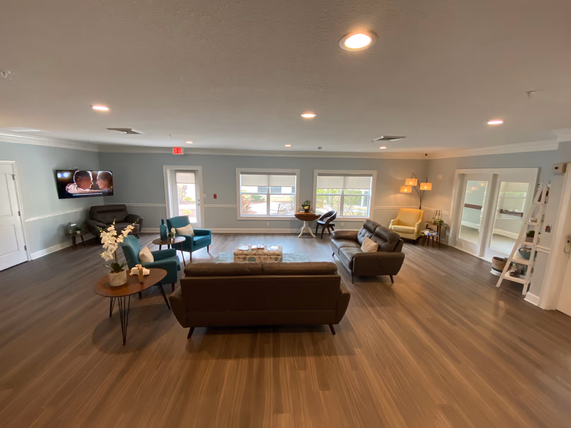 A spacious and well-lit living room area in Cypress Creek Assisted Living featuring modern furniture including two brown sofas, two teal armchairs, a yellow armchair, and a dark brown armchair. The room has large windows letting in natural light, a wall-mounted TV, wooden flooring, and various small tables with decorative items and plants.