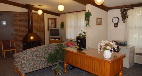A cozy living room area with a floral patterned couch facing a small TV on a wooden stand. There is a wooden desk with a computer monitor and a white vase with flowers on it. The room has a brick fireplace with a metal chimney, several hanging plants, framed pictures on the walls, and round ceiling lights. A wooden chair is placed near a door in the background.