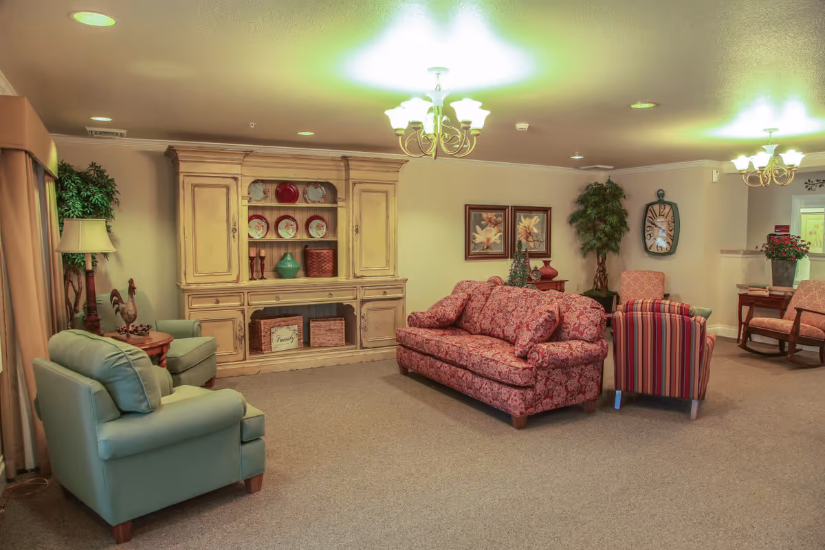 A cozy living room area with a patterned red sofa, a striped armchair, and two light blue armchairs. There is a large wooden cabinet with decorative plates and baskets, a floor lamp, potted plants, framed floral artwork on the wall, and a large wall clock. The room is softly lit by ceiling lights and chandeliers.