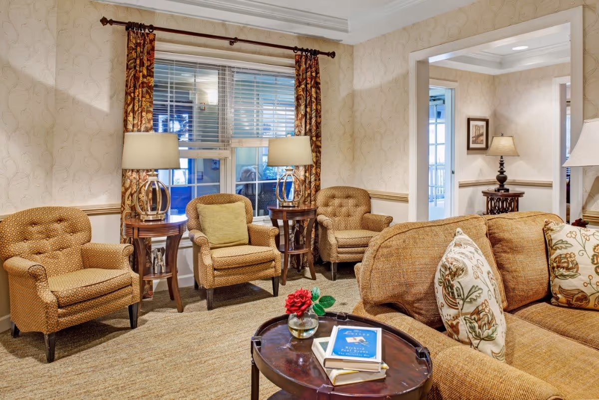 A cozy living room area with three patterned armchairs, two wooden side tables with lamps, a large window with floral curtains, and a brown sofa with floral cushions. A round wooden coffee table in front of the sofa holds a small vase with a red flower and a few books. The walls have a light patterned wallpaper, and there is an open doorway leading to another room with similar decor.