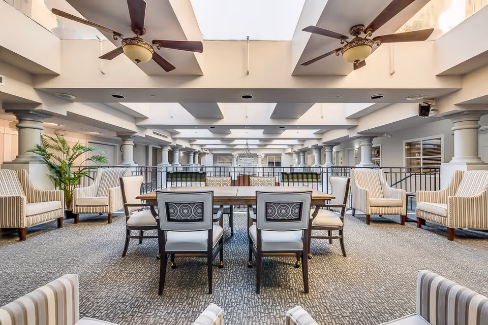 Spacious and well-lit senior living common area with a large wooden table surrounded by chairs in the center. The room features multiple striped armchairs arranged around the perimeter, ceiling fans, large skylights, and decorative columns along the sides.