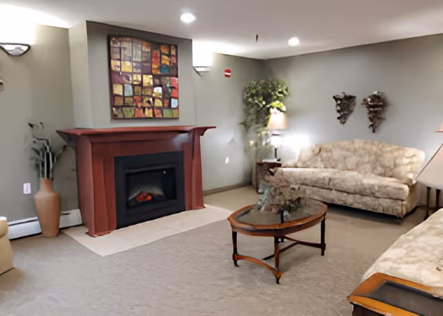 A cozy living room with a beige carpet, two floral-patterned sofas, a wooden coffee table with a glass top, a red fireplace with a colorful abstract painting above it, a tall potted plant, and two wall-mounted decorative pieces.