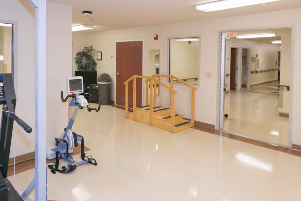 An indoor rehabilitation area with exercise equipment including a stationary bike and a treadmill. There is a wooden step exercise platform with handrails, a trash can, a potted plant, and a hallway leading to other rooms.