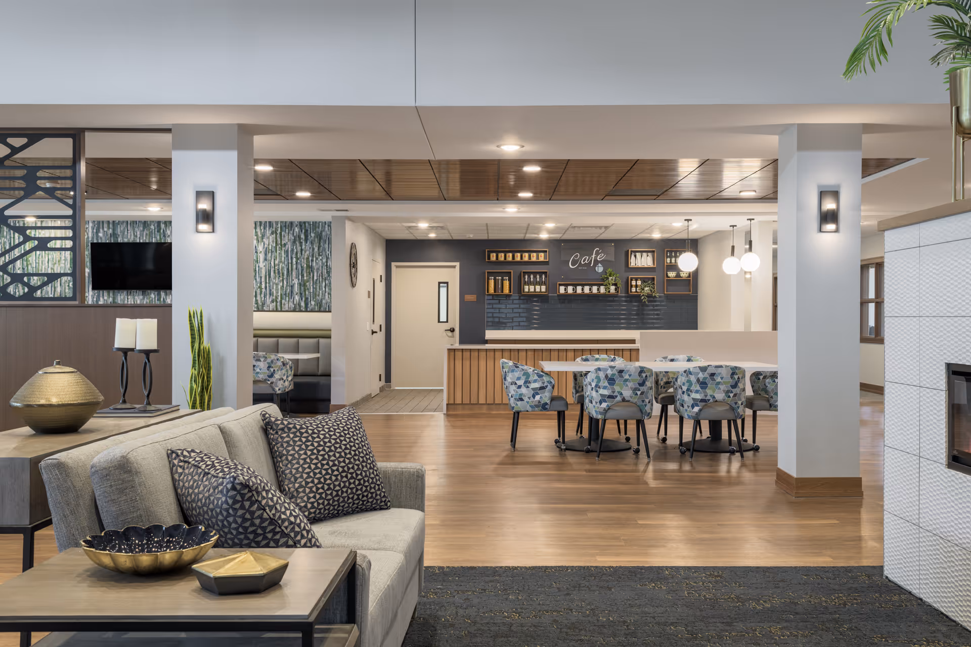 A modern senior living common area featuring a cozy seating area with a gray sofa and patterned pillows, a wooden coffee table with decorative bowls, and a dining area with a table and six patterned chairs. The back wall has a cafe counter with shelves displaying jars and plants, and the ceiling has recessed lighting with wooden paneling. There are white pillars and a fireplace on the right side.