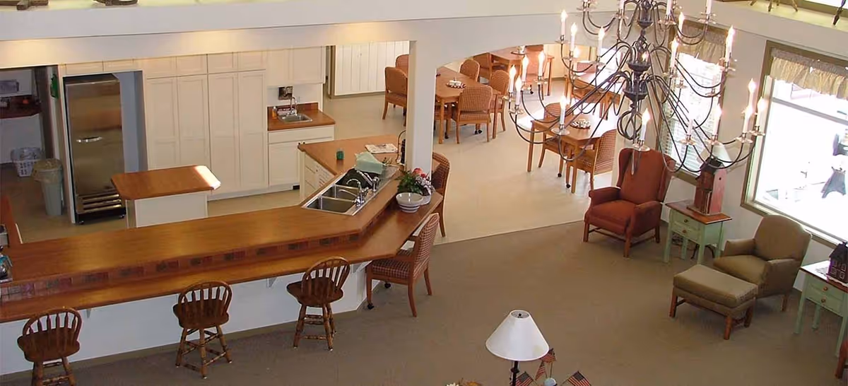 Interior view of a senior living facility showing a kitchen area with a wooden counter and three wooden chairs, a dining area with multiple tables and chairs, and a sitting area with armchairs and side tables near large windows. A large chandelier hangs from the ceiling.