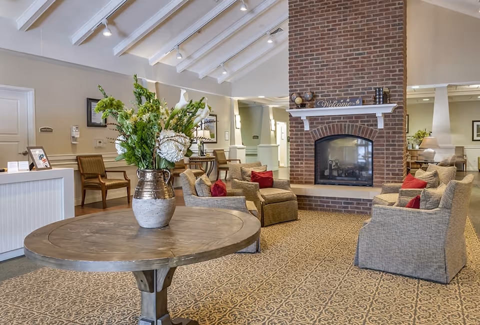 A cozy senior living facility common area with a round wooden table holding a large vase of green and white flowers in the foreground. In the background, there are several cushioned armchairs with red pillows arranged around a brick fireplace with a white mantel displaying decorative items and a 'Welcome' sign. The room has a patterned carpet, beige walls, and a high ceiling with exposed beams and recessed lighting.