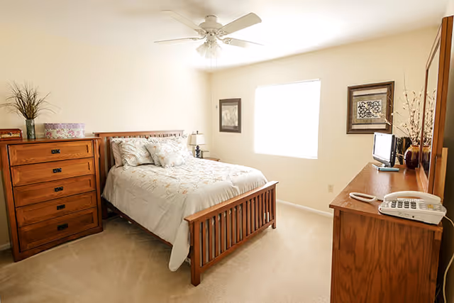 A bright bedroom with a wooden bed frame and a neatly made bed with floral bedding. There is a wooden dresser with a lamp, a small TV, a telephone, and decorative items on top. The room has beige walls, a window letting in natural light, and framed artwork on the walls.