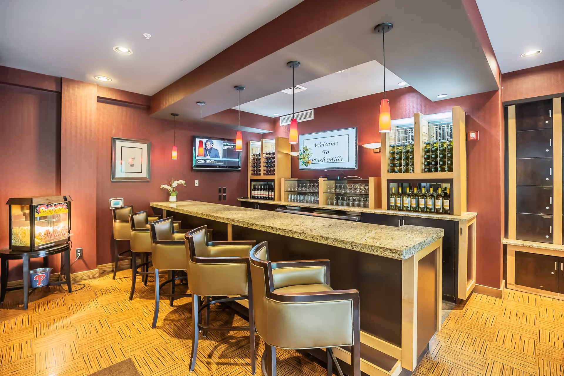 Interior view of a senior living facility bar area with a long granite countertop, several brown leather bar stools, wine bottles displayed on shelves, hanging pendant lights, a popcorn machine on a small table, and a wall-mounted TV. A sign on the wall reads 'Welcome To Plush Mills'.