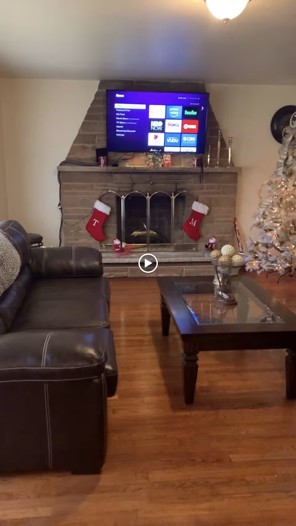 A cozy living room with a brown leather couch on the left, a wooden coffee table with decorative items in the center, and a brick fireplace with a mounted flat-screen TV above it. Two red Christmas stockings hang on the fireplace, and a decorated Christmas tree with white lights is visible on the right side.