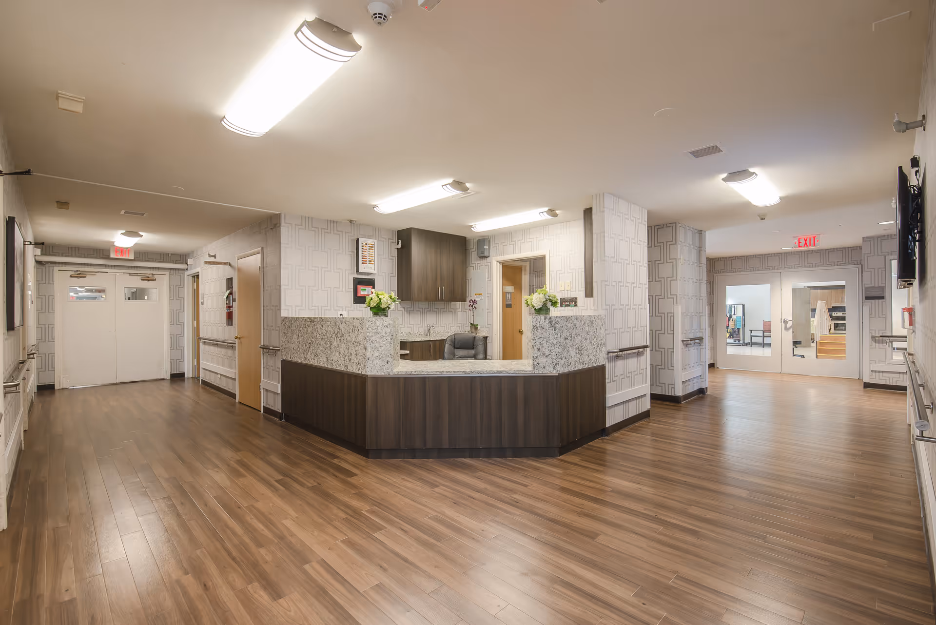 A spacious and well-lit reception area in a senior living facility with wood flooring, a large granite countertop desk, floral decorations, and multiple doors leading to other rooms. The walls have a patterned wallpaper, and there are ceiling lights and exit signs visible.