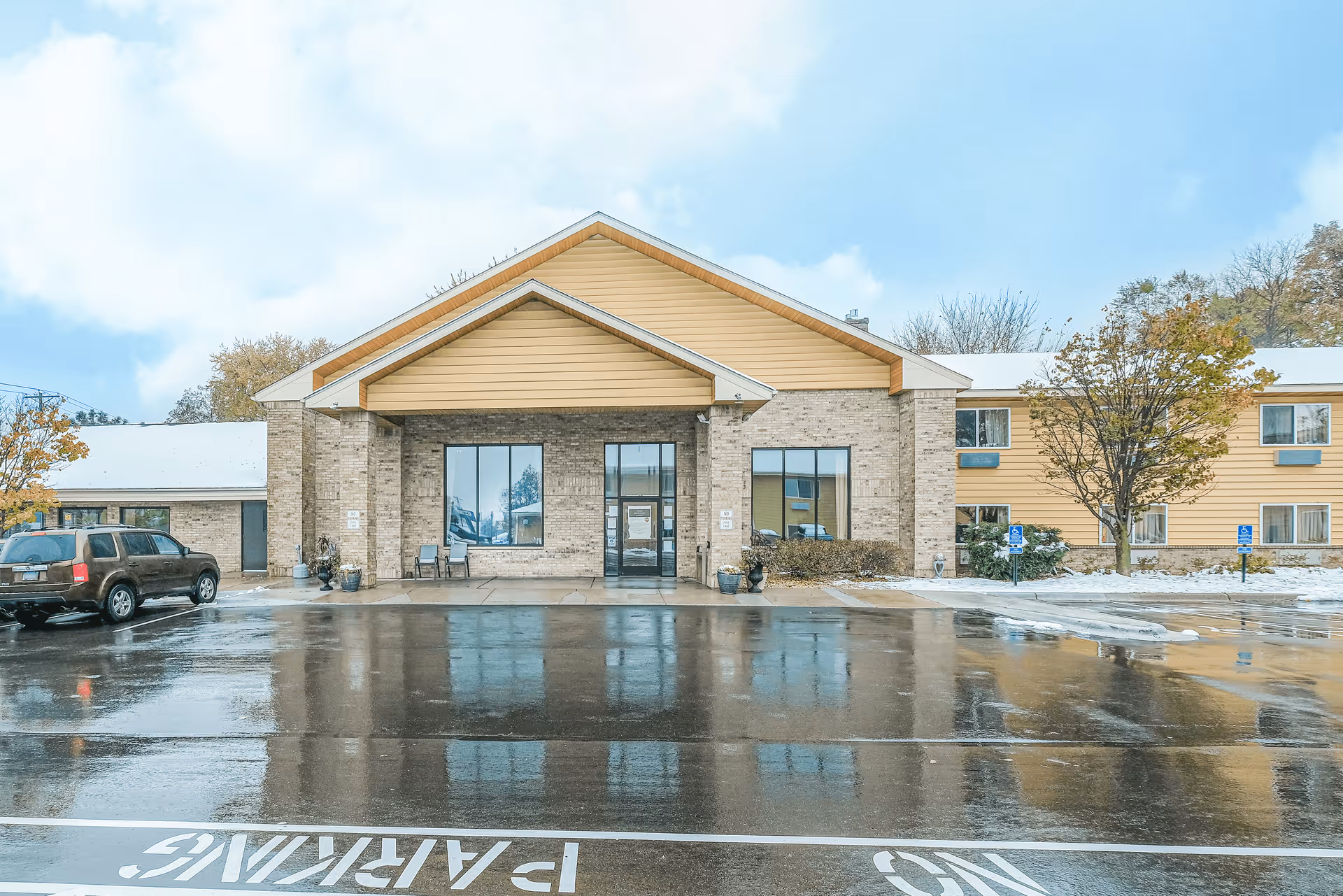 Front entrance of a two-story senior living building with a covered portico, wet reflective parking lot, and light snow on the ground.