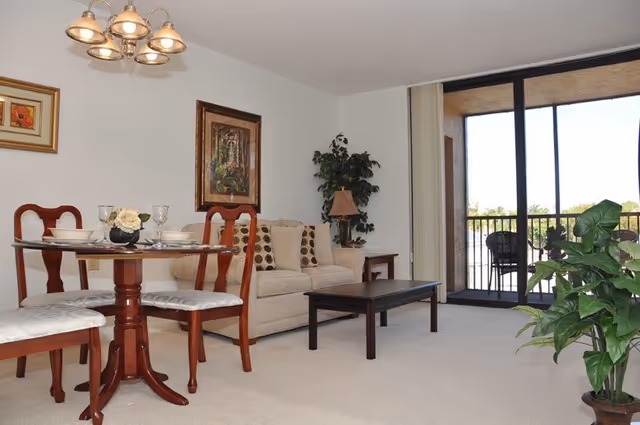A living room and dining area with a beige sofa adorned with patterned cushions, a wooden coffee table, and a round dining table with four chairs. The room has framed artwork on the walls, a potted plant, a lamp on a side table, and a sliding glass door leading to a balcony with outdoor seating and a view of greenery.