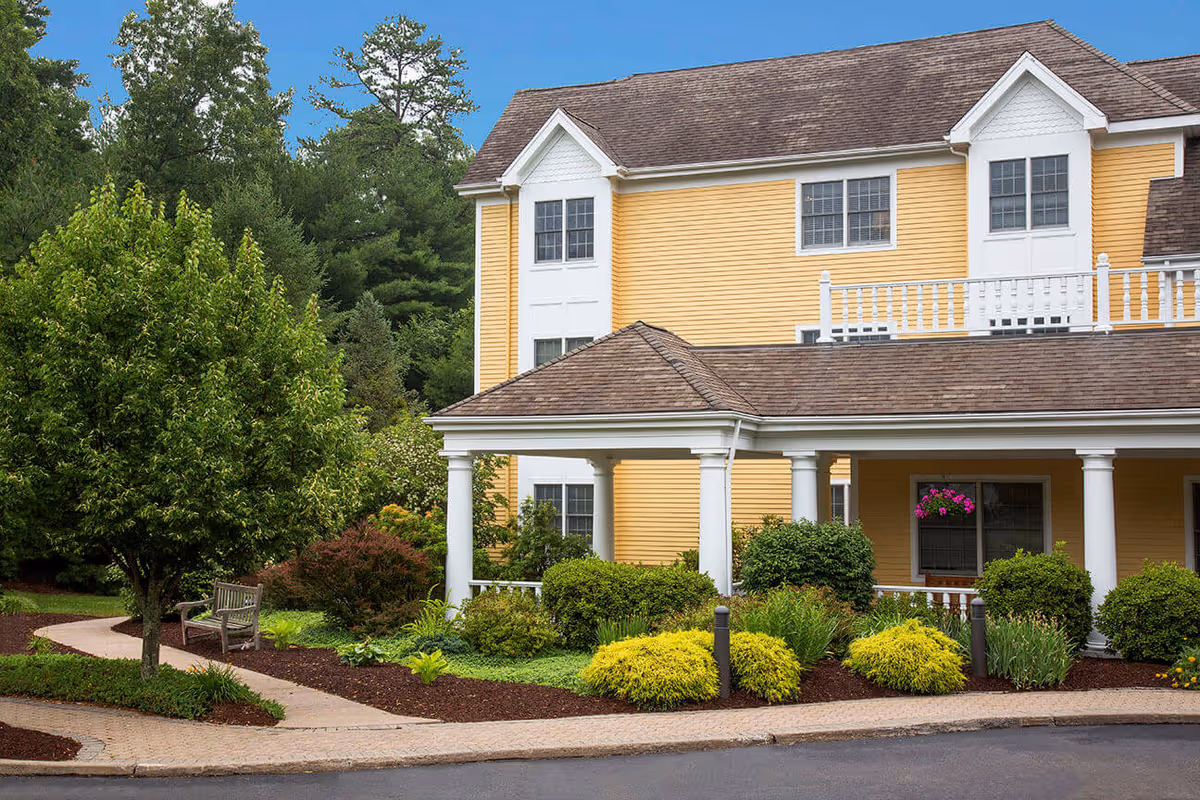 Exterior view of a yellow two-story building with white trim and a covered porch, surrounded by green trees, bushes, and landscaped garden beds with a paved walkway and a wooden bench.