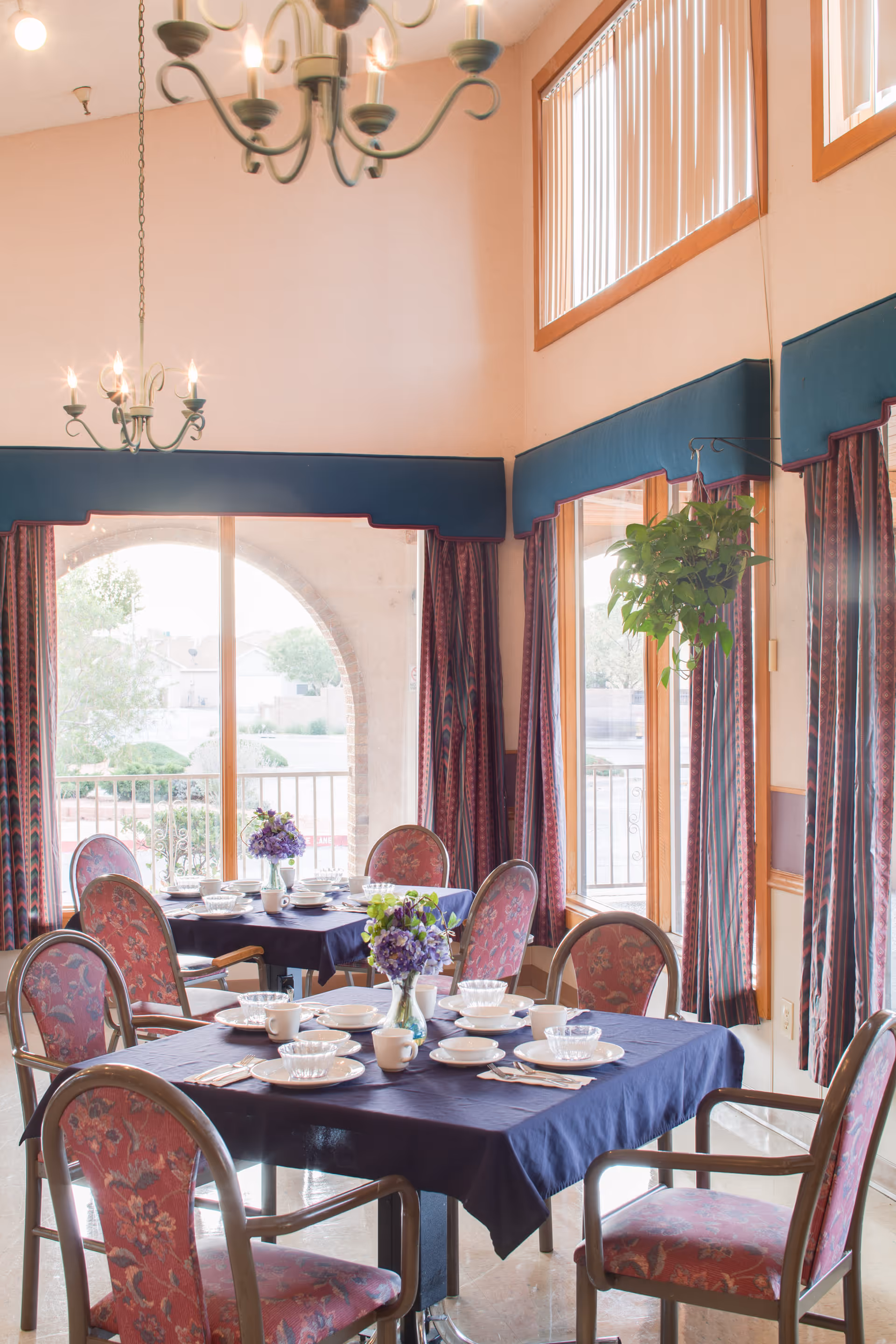Dining room with two tables covered in dark blue tablecloths, each set with white plates, bowls, cups, and silverware. The chairs have red floral upholstery. There are two chandeliers hanging from the ceiling, large windows with vertical blinds and patterned curtains, and a hanging green plant near the window.