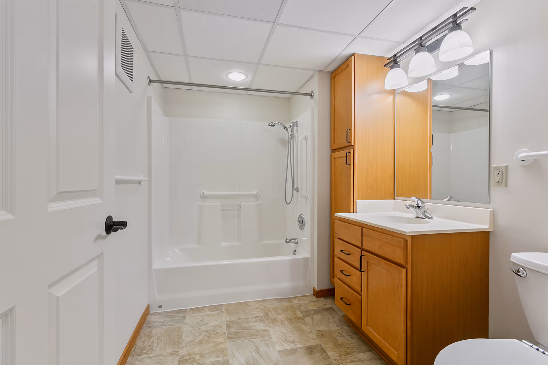 A clean and well-lit bathroom featuring a white bathtub with a showerhead and grab bars, a wooden vanity with a sink and mirror above, a toilet, and tiled flooring. The walls are light-colored and there is a white door partially open on the left side.