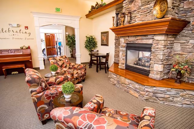 A cozy living room area with a stone fireplace and wooden mantel. Three floral-patterned armchairs are arranged around a small round table with a potted plant. In the background, there is a wooden piano against the wall and a small table with two chairs near a doorway. The walls are light-colored, and there is a decorative sign on the wall that reads 'Celebrating'.