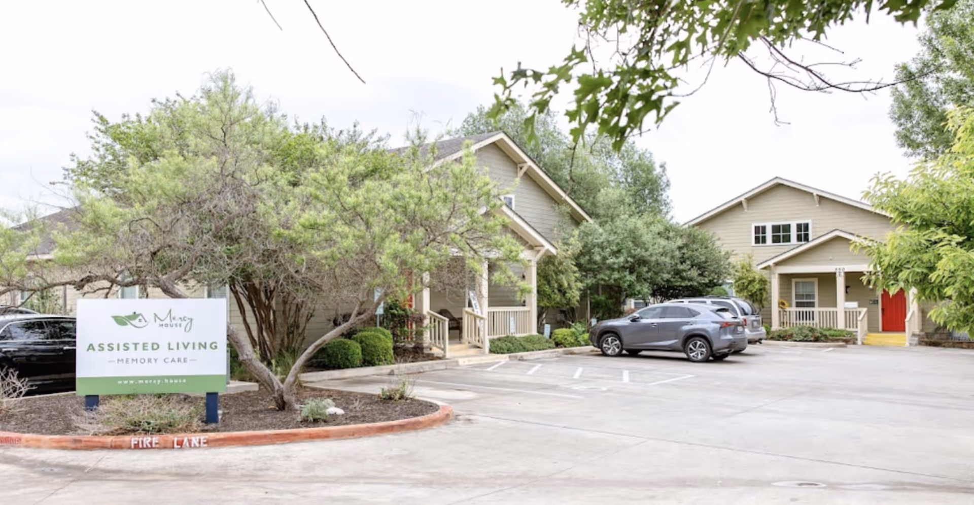 Front exterior of Mercy House assisted living facility showing the entrance buildings, parking area, landscaping, and a sign reading 'Assisted Living Memory Care'.