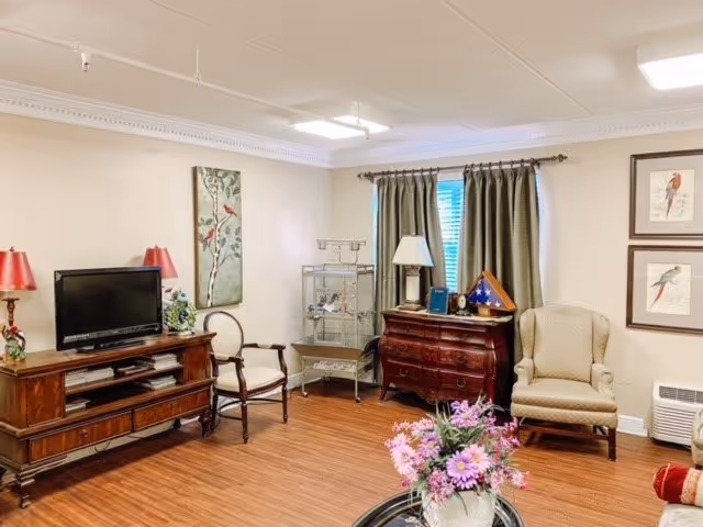 A cozy living room area in Atmore Nursing Center featuring a wooden TV stand with a flat-screen TV and two red lamps, a wooden chair with white upholstery, a birdcage, a wooden dresser with a lamp and folded American flag, an armchair, framed bird artwork on the wall, a window with green curtains, and a round table with a vase of pink flowers in the foreground.