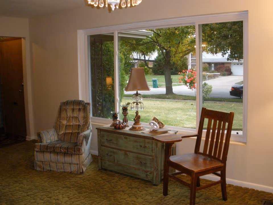 A cozy interior room with a large window showing a view of a green lawn and trees outside. Inside, there is a striped upholstered armchair on the left, a wooden chair on the right, and a rustic green chest of drawers in the center with a decorative lamp and small ornaments on top. The floor is covered with a green patterned carpet.