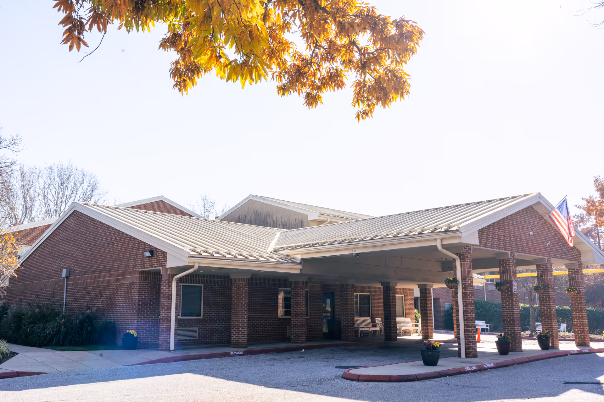 Front of a brick building with a covered drop-off porte-cochère, potted plants, seating, and an American flag under autumn foliage.