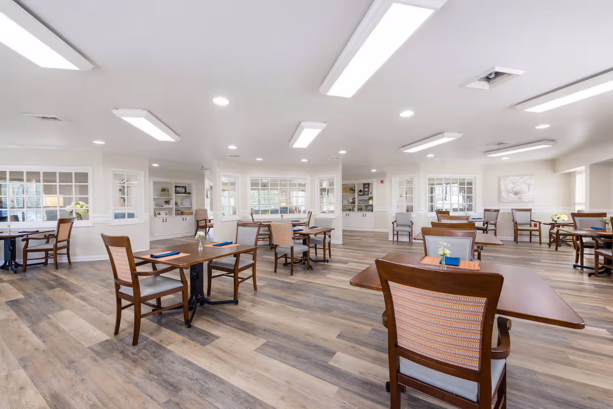 A spacious and well-lit dining room with multiple wooden tables and chairs arranged neatly. The room features large windows, built-in shelves with decorative items, and light wood flooring. The walls are painted white, and there are ceiling lights providing bright illumination.