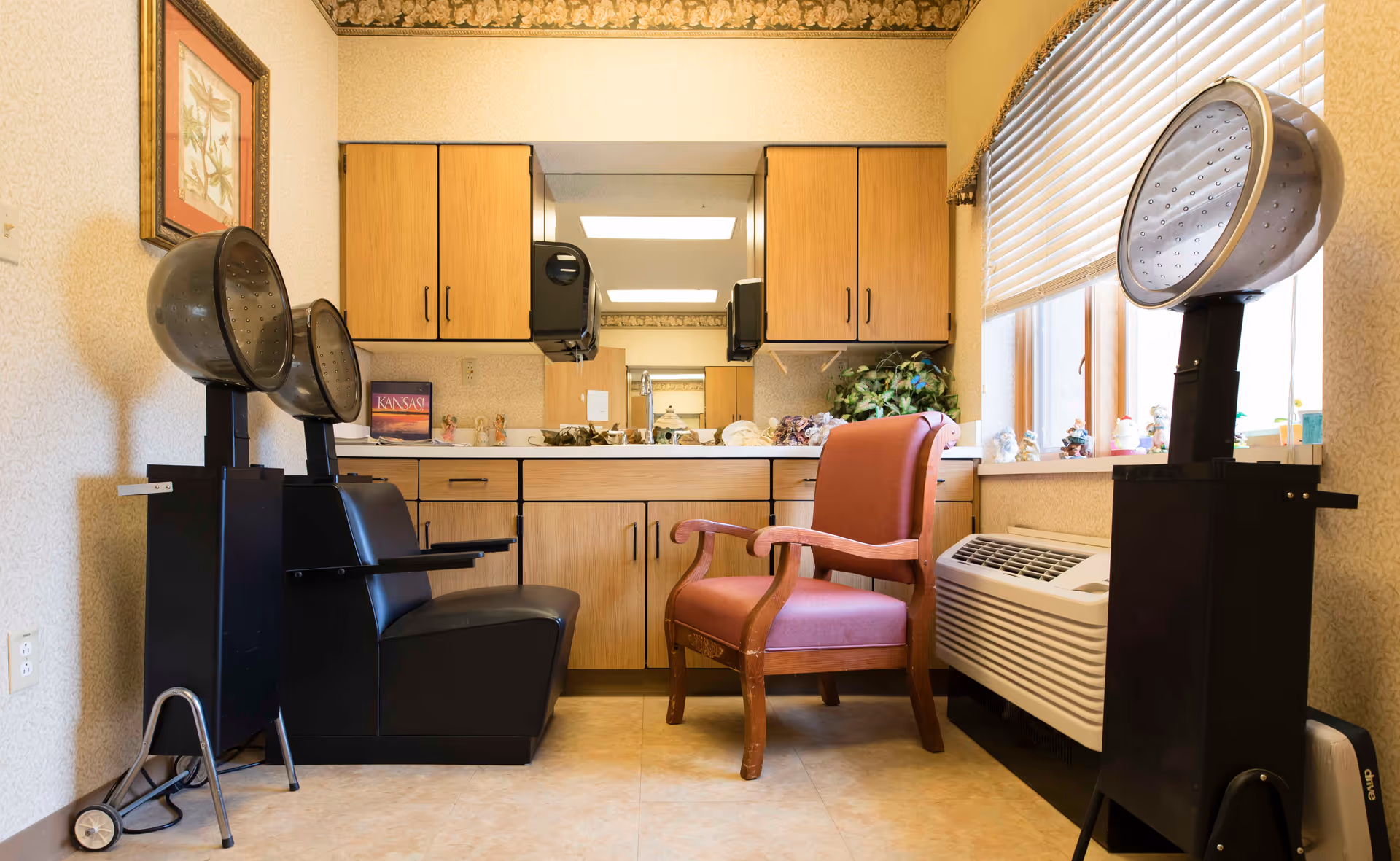 Interior view of a hair salon area in a senior living facility with two black hair drying chairs on the left and right sides, a pink cushioned wooden chair in the center, wooden cabinets with a mirror above the counter, and a window with blinds on the right side.