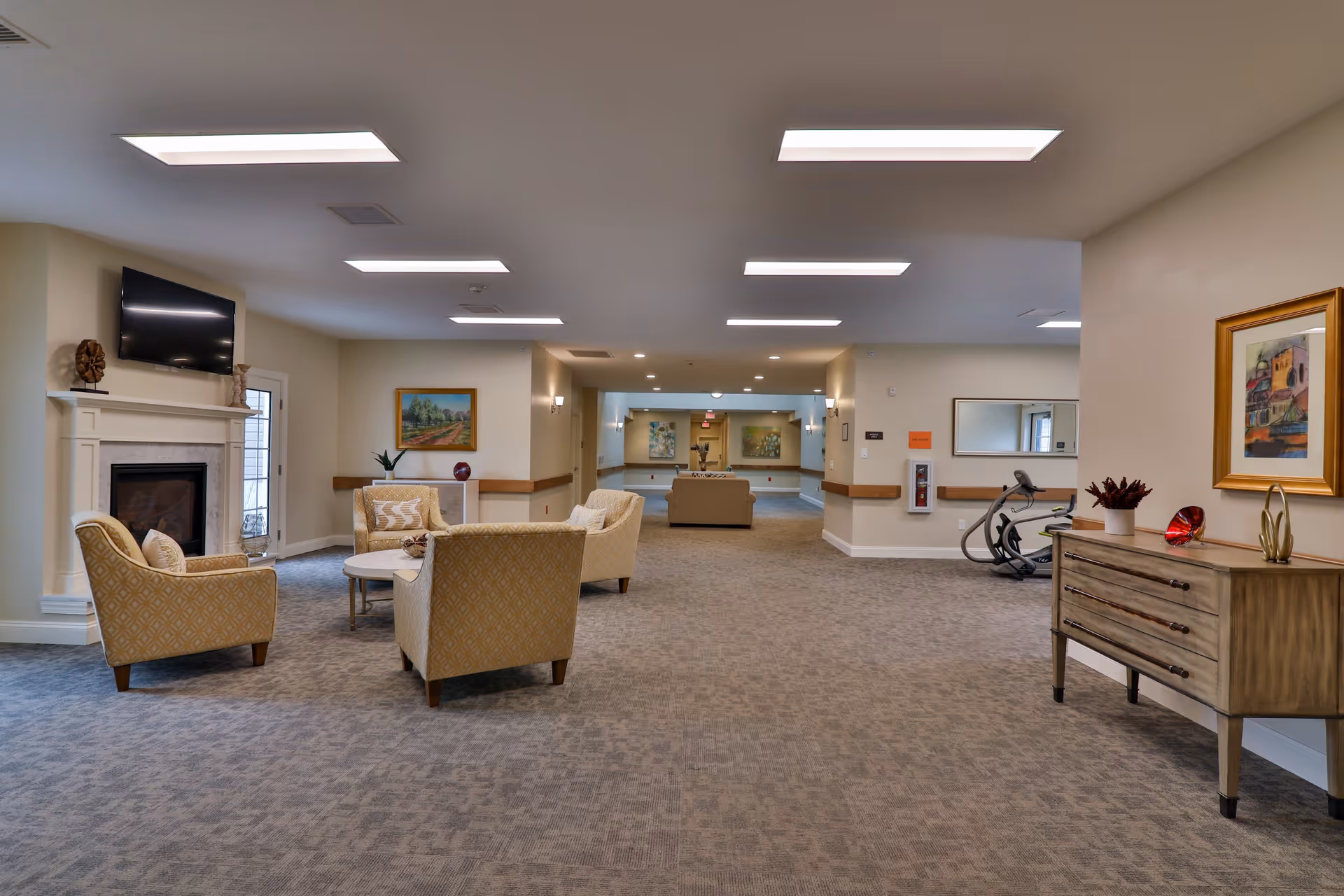 Spacious senior living facility common area with beige armchairs arranged around a small round table near a fireplace with a mounted TV above it. The room has carpeted floors, neutral-colored walls, ceiling lights, framed artwork, and a wooden dresser with decorative items. An exercise bike is visible in the background.